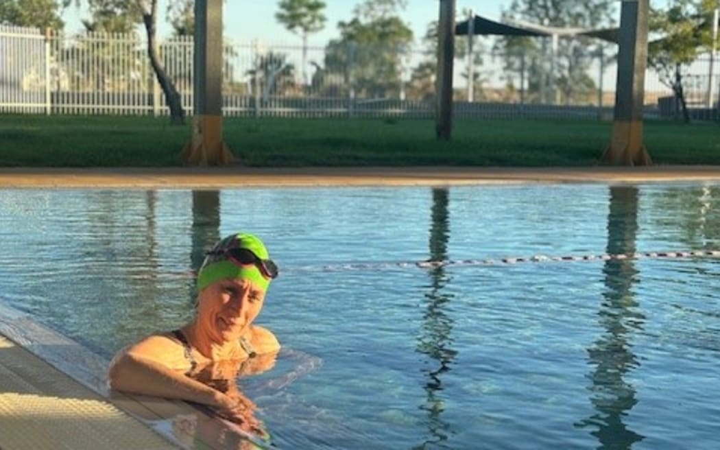 Janene is in an outdoor lap pool with a swimming cap and goggles on.