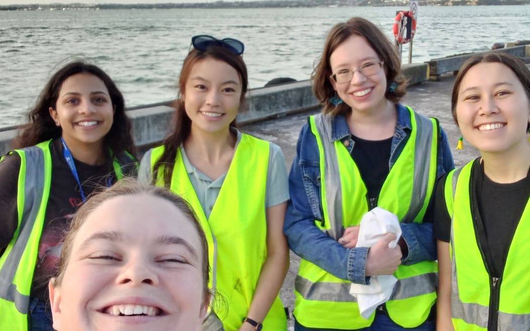 A selfie featuring four women in high-vis vests standing in front of the water. The face of the woman taking the selfie is visible in the foreground. Everyone is smiling.