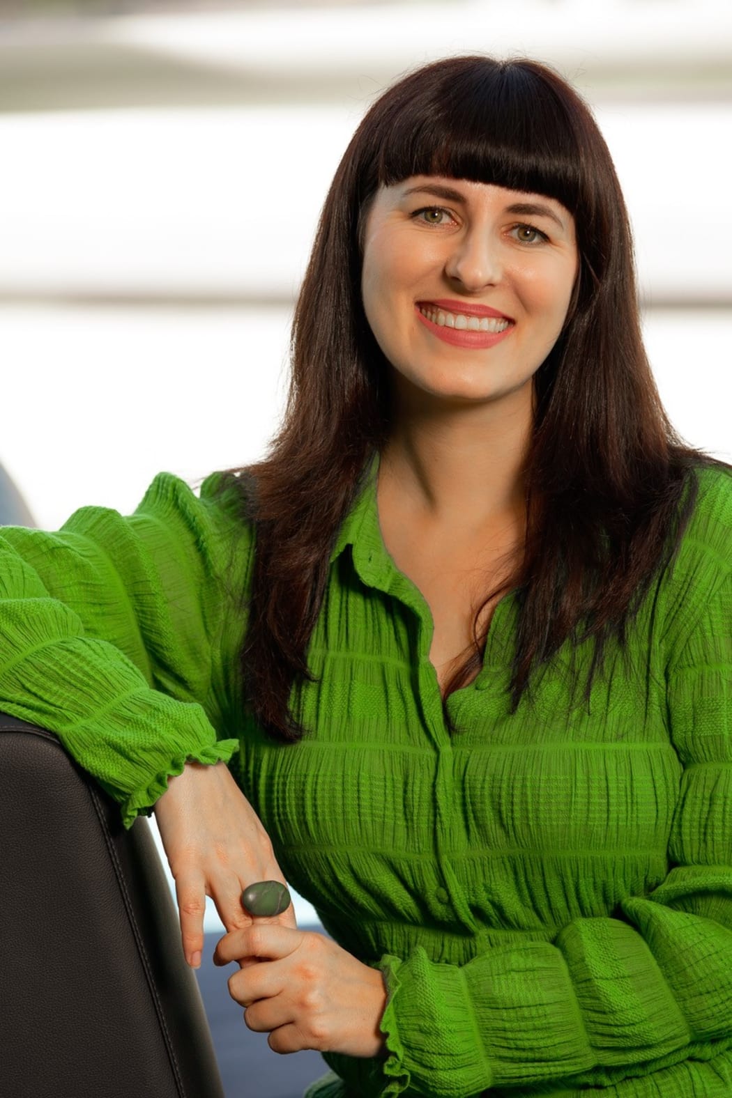 Jane sits on a chair and smiles at the camera. She wears a vibrant green top and a large green ring.