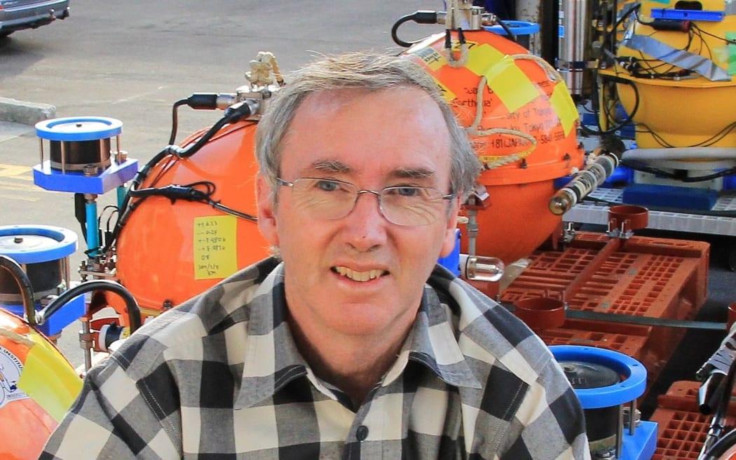 A man in a black and white checked shirt and glasses smiles at the camera while sitting in front of orange cylinder devices.
