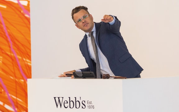 Charles Ninow is conducting an auction. He is looking somewhere behind the camera and gesturing with his hand. He is standing behind a white podium with "Webb's - Est. 1976" written on it in black lettering. To his right is the side of a large, colourful painting.