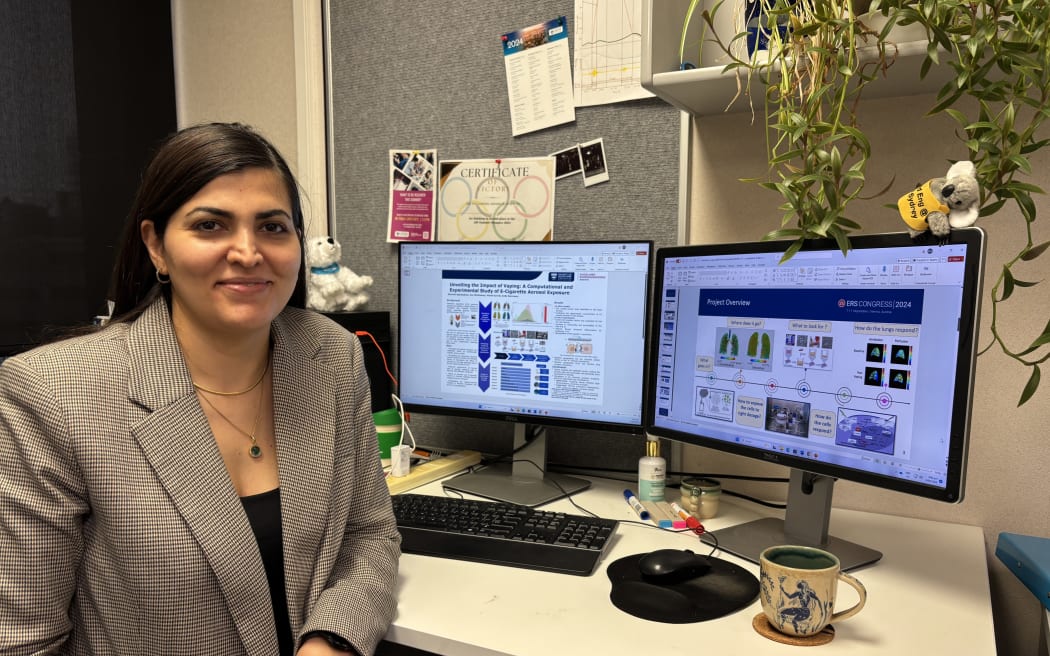 A woman in a grey blazer sits at a desk in front of two monitor screens. She is turned towards the camera and smiling.