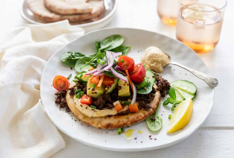 Spiced lamb mince with hummus and flatbreads on a round white plate garnished with herbs and a lemon wedge.
