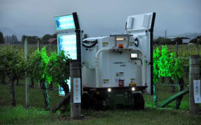 A robot with a UV-light panel attached to its side, drives down a row of vines.