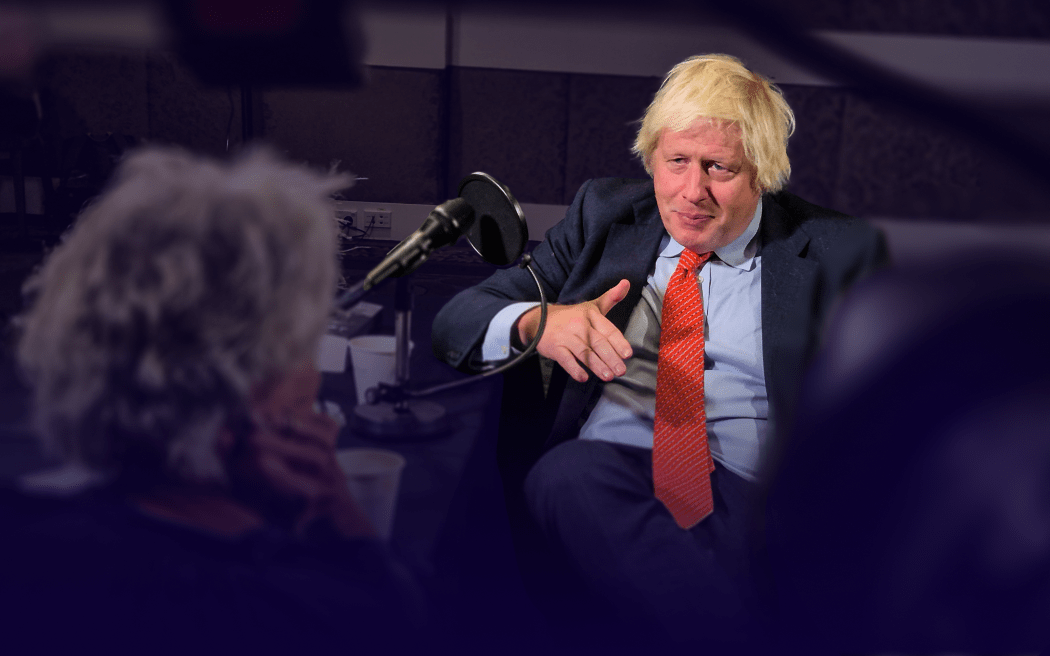 A man with floppy blonde hair wearing a suit gestures while talking in a studio.