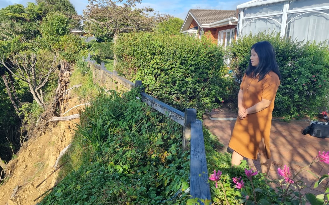 Jennifer Chen has black long hair and wears an orange dress. She stands at the edge of her property looking down at the cliff which has fallen away.