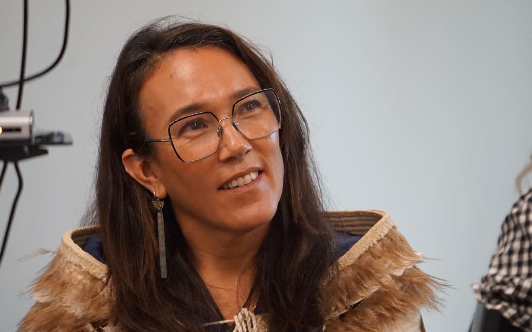 Claire Charters is wearing glasses with long brown hair, she is sitting down and is looking upwards, as if she is listening.