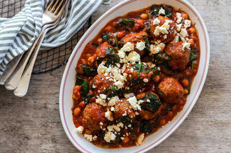 An oval dish on a wooden board containing Spanish meatballs with chilli, chickpea and spinach sauce