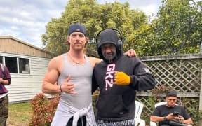 Two men standing on a lawn in what appears to be a back garden. There is a fence and a white garage in the background.