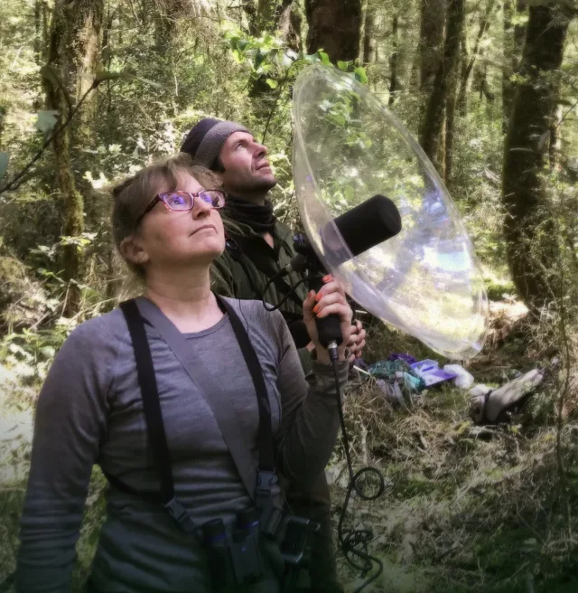 A woman with binoculars around her neck holds up a large plastic dish surrounding a microphone in the forest. Behind her, a man in a beanie looks up towards the canopy.