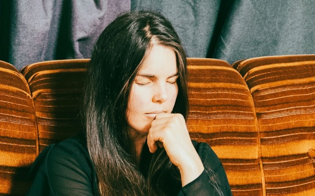 Christchurch musician Mel Parsons, a white woman with long dark hair, sits on a brown couch with her eyes closed and a hand up to her mouth