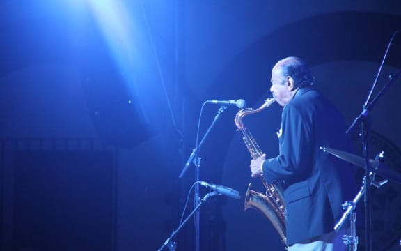 Saxophonist playing into a microphone, suffused in blue light.