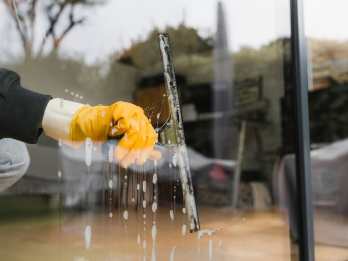 A hand in a yellow plastic glove cleans a window with a squeegee.