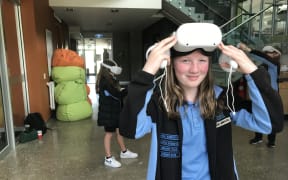 A student lifts up a virtual reality headset so you can see her face. She is holding two controllers. Behind her are two other students with headsets on.