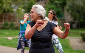 A white-haired woman in a black t-shirt raises her arms in an outdoor exercise class.