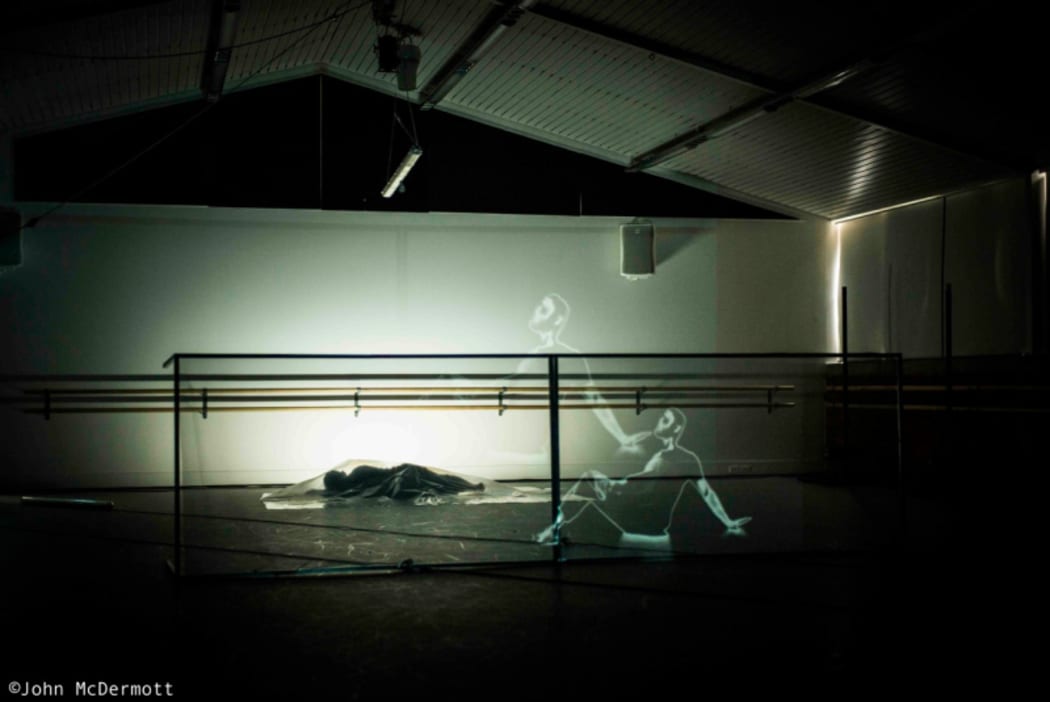 A production still from In Transit showing a dancer lying behind a clear screen on which an image of another dancer is being projected.