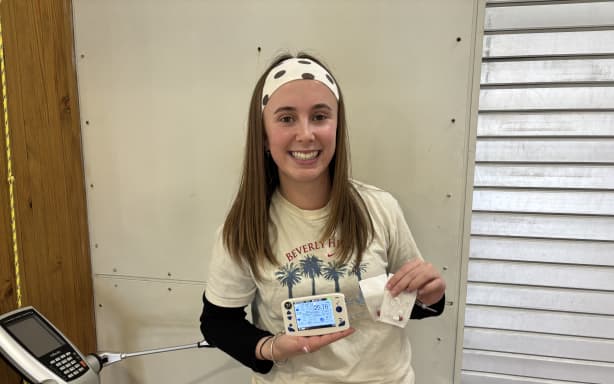 Riley is standing next to a body fat and muscle mass machine, which looks slightly like a smaller version of an exercise machine. She is holding a small monitor in her right hand, pointed out towards the camera, and some plastic wrapped core temperature logging devices in her left. They look like small red and white pills. She is smiling, looking at the camera, with a polka dot head band, long brown hair and a long sleeved black top with a beige t-shirt over it.
