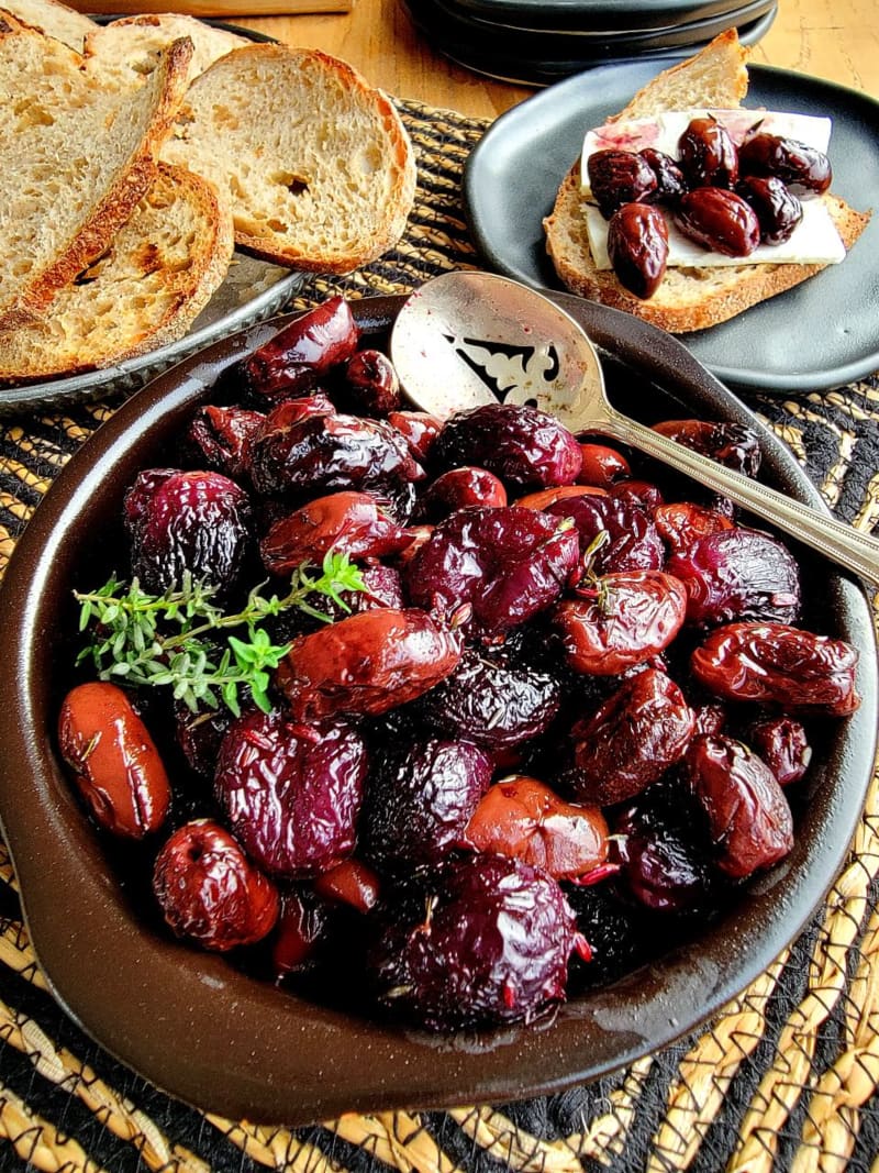 Roasted red grapes and Kalamata olives on a plate near two plates with bread and feta cheese.