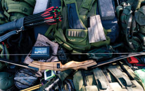 A pile of prepper gear: military vests, rucksacks, a bow and a quiver of arrows.