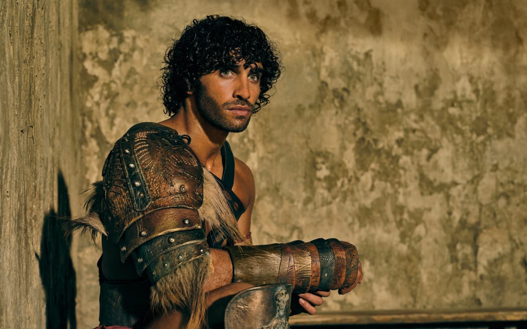 Jordi Webber, a blue-eyed man with dark, curly hair, wears Roman armou and sits against a brown wall.