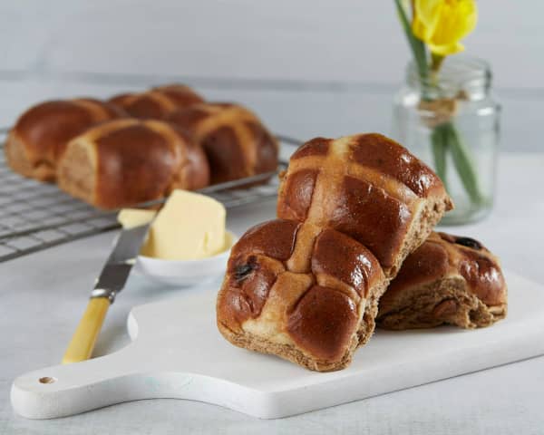 A pair of hot cross buns are stacked up on top of a single bun, with more in the background.