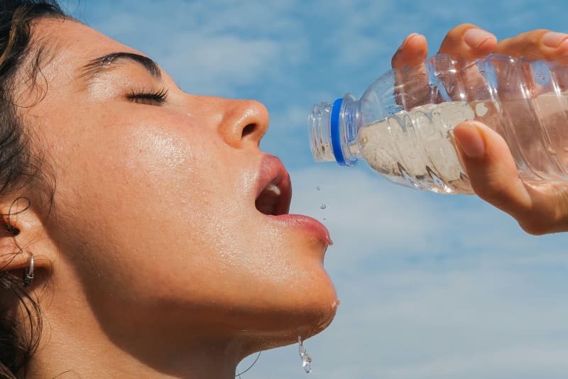 A woman opens her mouth to drink from a water bottle in the sun outdoors.