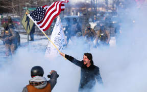 A man waves a US flag next to people protesting US Immigration and Customs Enforcement (ICE) near the Bishop Henry Whipple Federal Building in Minneapolis, Minnesota, on January 9, 2026. A US Immigration and Customs Enforcement (ICE) agent shot and killed an American woman on the streets of Minneapolis January 7, leading to huge protests and outrage from local leaders who rejected White House claims she was a domestic terrorist. The woman, identified in local media as 37-year-old Renee Nicole Good, was hit at point blank range as she apparently tried to drive away from agents who were crowding around her car, which they said was blocking their way. (Photo by CHARLY TRIBALLEAU / AFP)
