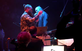 Musicians Jeremy Mayall and Horomoa Horo perform on stage. Jeremy is conducting and holds a baton, Horomona is playing taonga puoro.