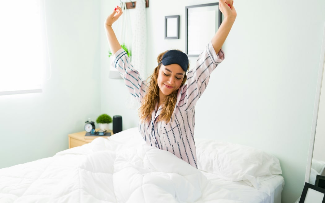 A woman wearing pyjamas and an eye mask gets up from bed stretching.