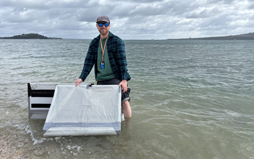 Tom Dowling stands in shallow water at Mission Bay Beach with his pant legs rolled up, holding a prototype of the SAR4SaR radar reflector made from corflute, soft plastic and duct tape.