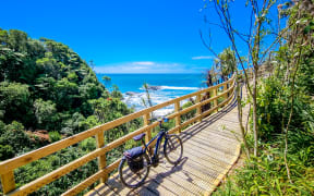 A bike leans against a fence on a newly constructed boardwalk. The boardwalk is on a gradual incline. It's coming out of a patch of native bush, you can see the sea emerging on the left hand side.