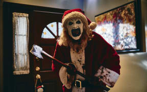 A still from the movie Terrifier. Someone dressed in a Santa suit with black and white face paint, covered in blood, walks through a hallway carrying an axe.