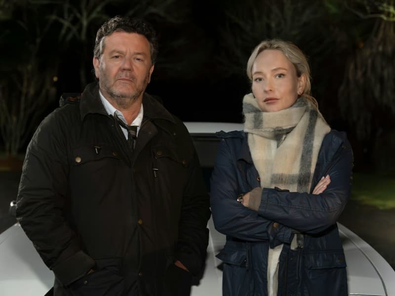 A man and a woman wearing coats give serious looks to the camera while standing in front of a car.