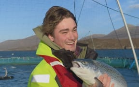 James Sibley is stood by a fish farm. He is holding a fish which looks a bit like a salmon.