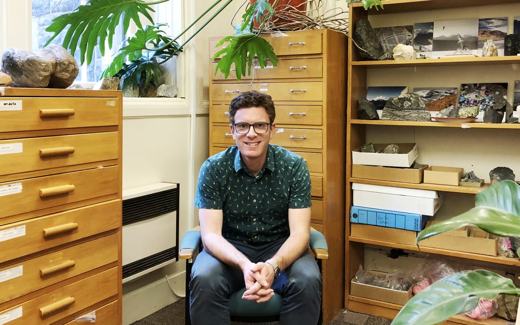 James Scott is sitting on a chair in an office surrounded by specimen drawers and rock samples on shelves.