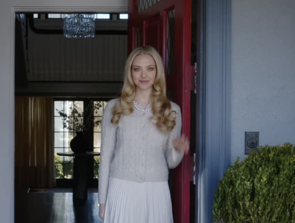 Amanda Seyfried, with styled blonde hair and wearing a grey cardigan, stands in front of an open front door.