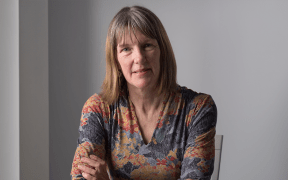 Kathleen folds her arms and smiles at the camera. She sits at a table.