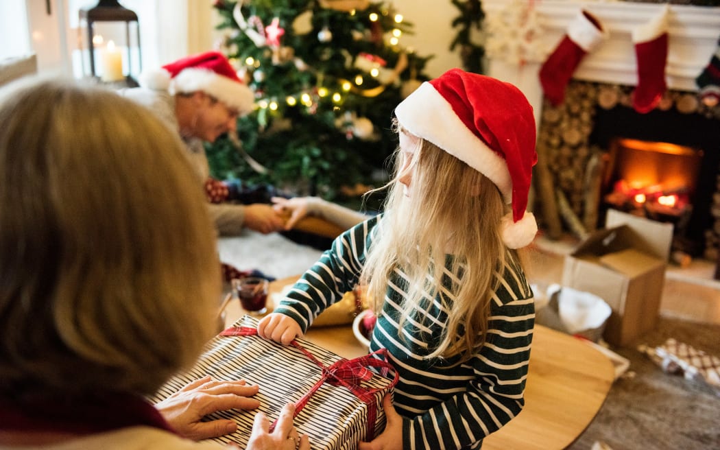 A young girl wearing a Santa hat is opening up a present, held by an adult, in a lounge with a Christmas tree.