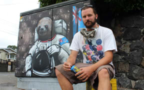 Paul sits beside a panting on a streetside utility box. He's painted a sloth wearing a spacesuit beside a New Zealand flag.