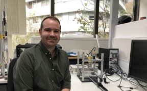 James sits next to a bench on which you can see a metal frame with an injector mounted vertically, a perspex box underneath into which skin is placed, and a track opposite this with a camera on it.