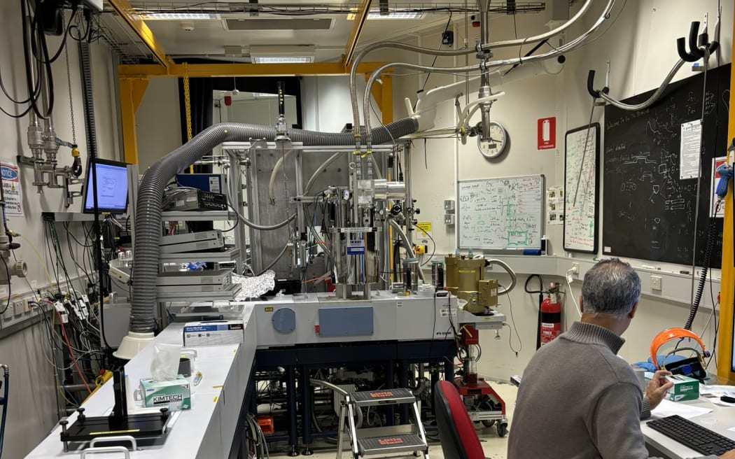On the right of the image, a man sits at a computer desk, he is looking away from the camera towards a large piece of complex metal scientific equipment, sitting on a bench in the centre of the image, with lots of pipes extending from it. The room is a lab with an industrial feel.