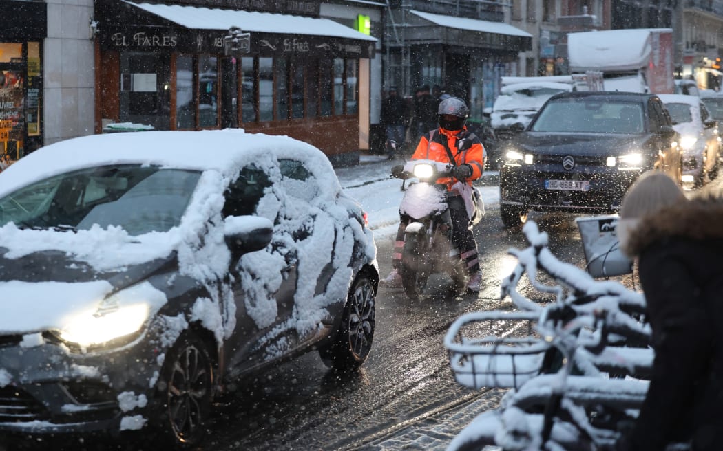 A diver steers a scooter next to cars on a road during a snowfall in Paris on January 5, 2026. (Photo by Ludovic MARIN / AFP)