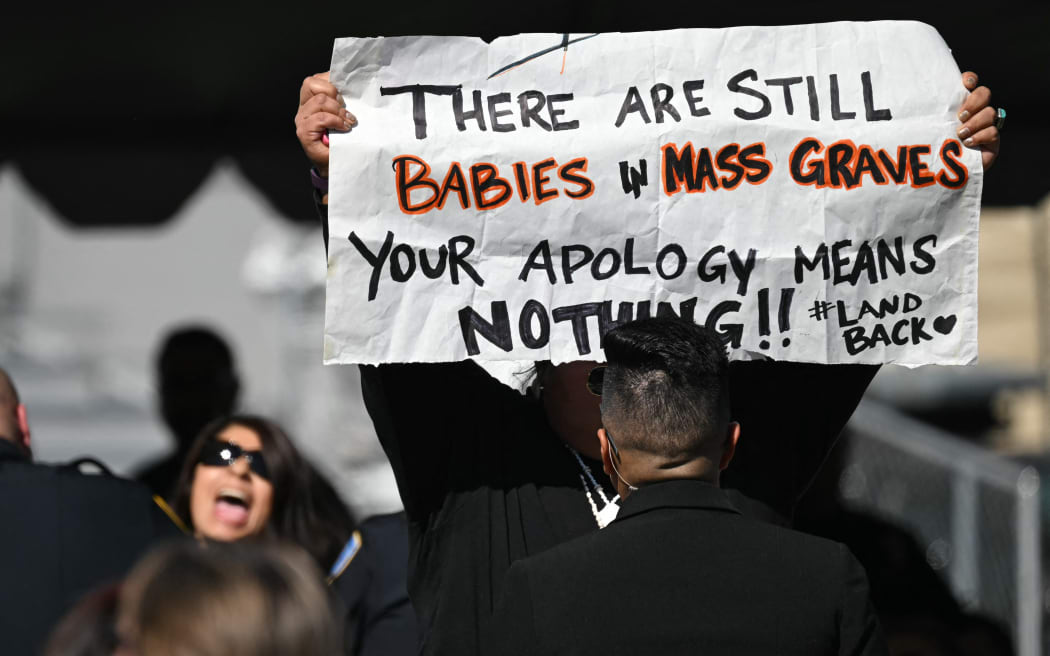 A protester holds a sign as US President Joe Biden speaks at the Gila River Crossing School in the Gila River Indian Community, in Laveen Village, near Phoenix, Arizona on October 25, 2024. - Biden apologized for one of the country's "darkest chapters:" the abduction of Native American children from their families and placement in abusive boarding schools aimed at erasing their culture. The first public apology issued by a sitting US president. From the early 1800s until the 1970s, the US ran hundreds of Indian boarding schools across the country to forcibly assimilate Native children into European settler culture, including conversion to Christianity. (Photo by ANDREW CABALLERO-REYNOLDS / AFP)