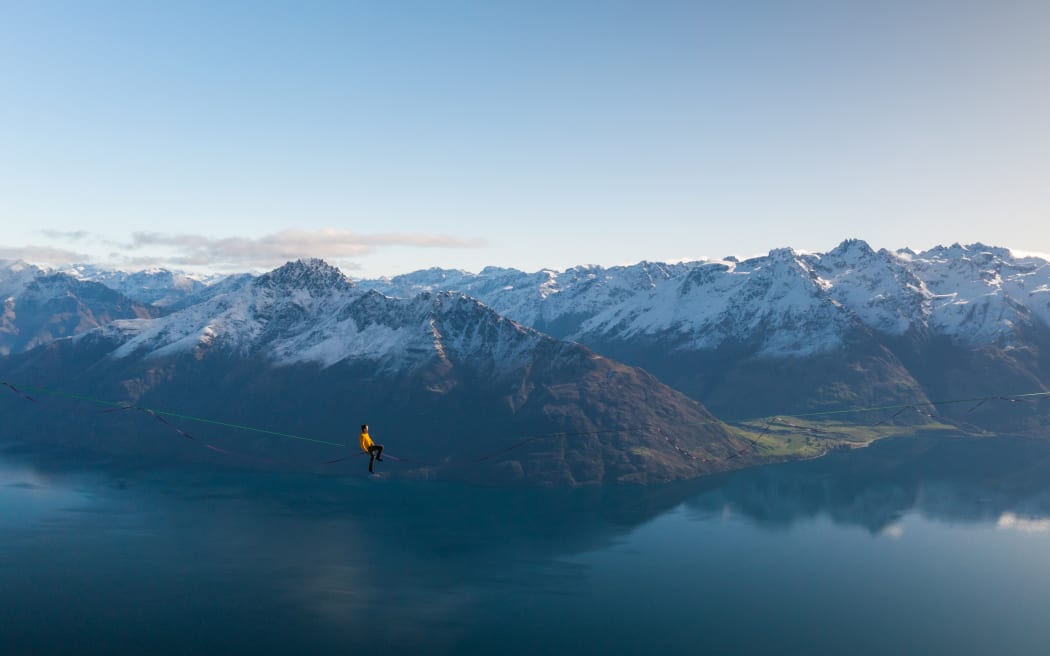 Gavin Alexander Lewis on a highline with the Remarkables in the background. The photo was taken by Will Nelson and is used by RNZ with permission. Will is crouched in the snow on a small ledge at 1400 metres, operating a drone to take the photo.