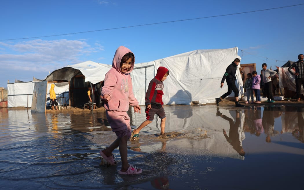 Displaced Palestinian children walk through a puddle of rain water at a makeshift camp housing displaced Palestinians in Khan Yunis, in the southern Gaza Strip on January 1, 2025, amid the continuing war between Israel and the militant Hamas group. (Photo by BASHAR TALEB / AFP)
