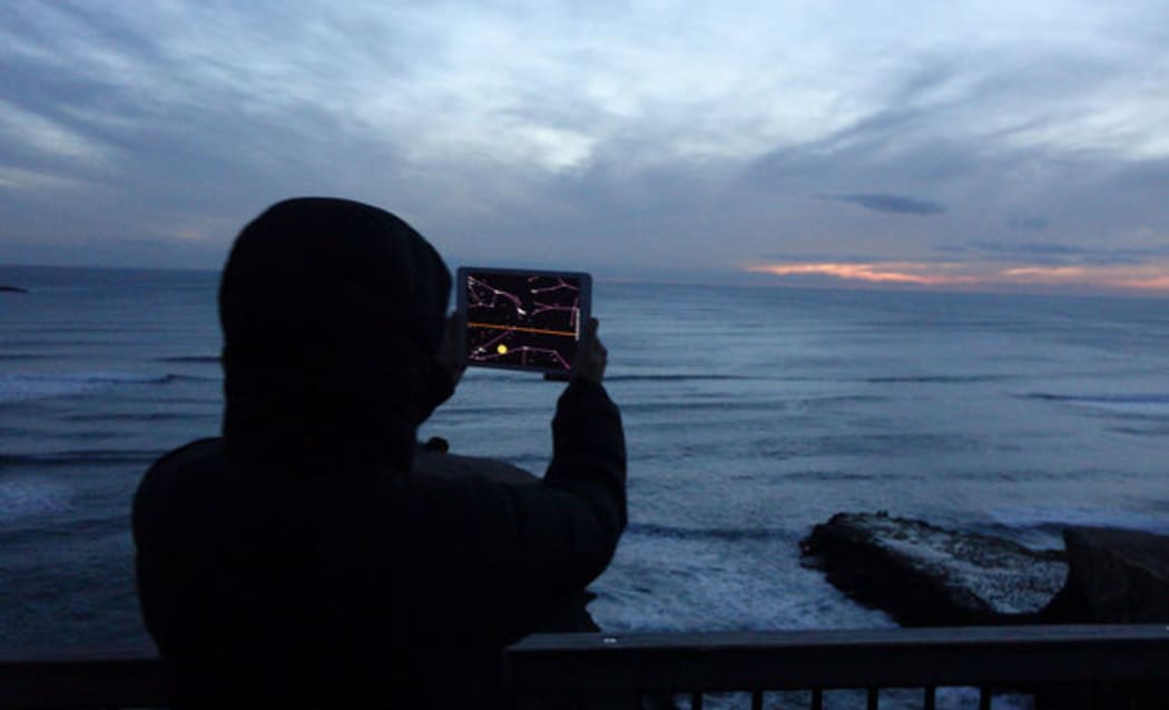 The group use an IPad to pinpoint the location of the rising moon among the clouds at Māori Bay at Muriwai Beach, Auckland.