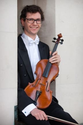 Alexander McFarlane is a viola player with the New Zealand Symphony Orchestra. He is wearing black and white concert attire, and holds his viola on his knee.