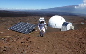Sheyna Gifford walking outside her simulated Mars base in a spacesuit. The base is an white dome and there are solar panels in front of it.