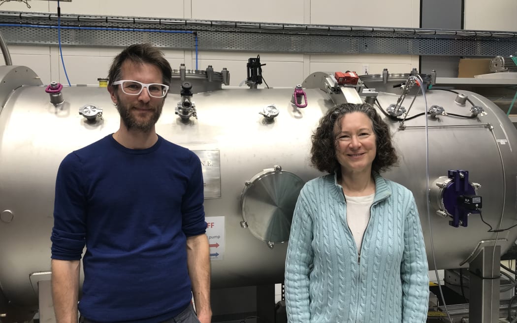 A man and a woman stand in front of a large metallic cylinder with various valves in a lab.