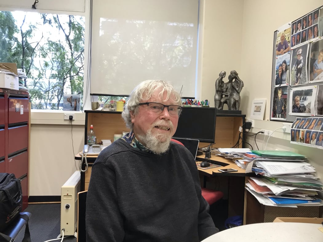 Warren Tate sitting at a desk in his office with photos on the wall and files on the top of shelves and tables behind him.